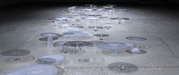 Glass discs sit atop oblong glass vessels on a stone floor. They are lit from above.