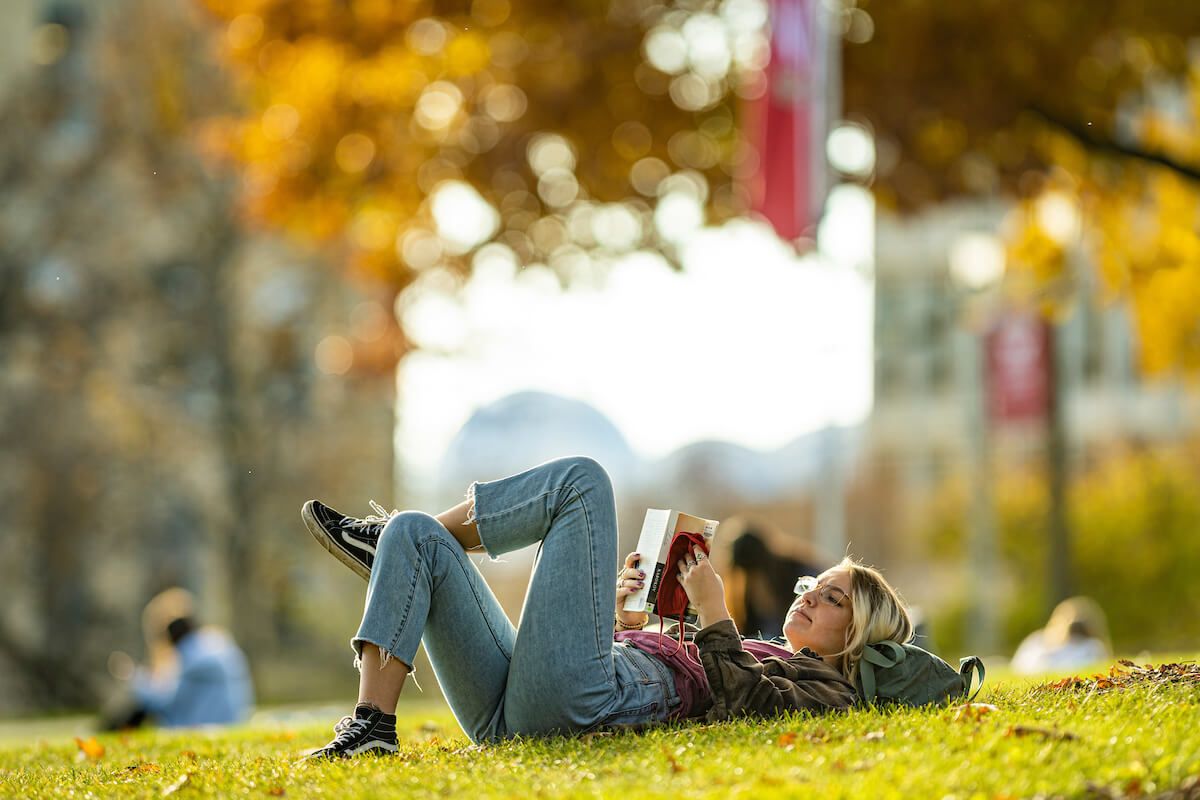 A young woman lays on Bascom Hill with one leg crossed over the other, reading a book. The background includes fall foliage and a UW banner.