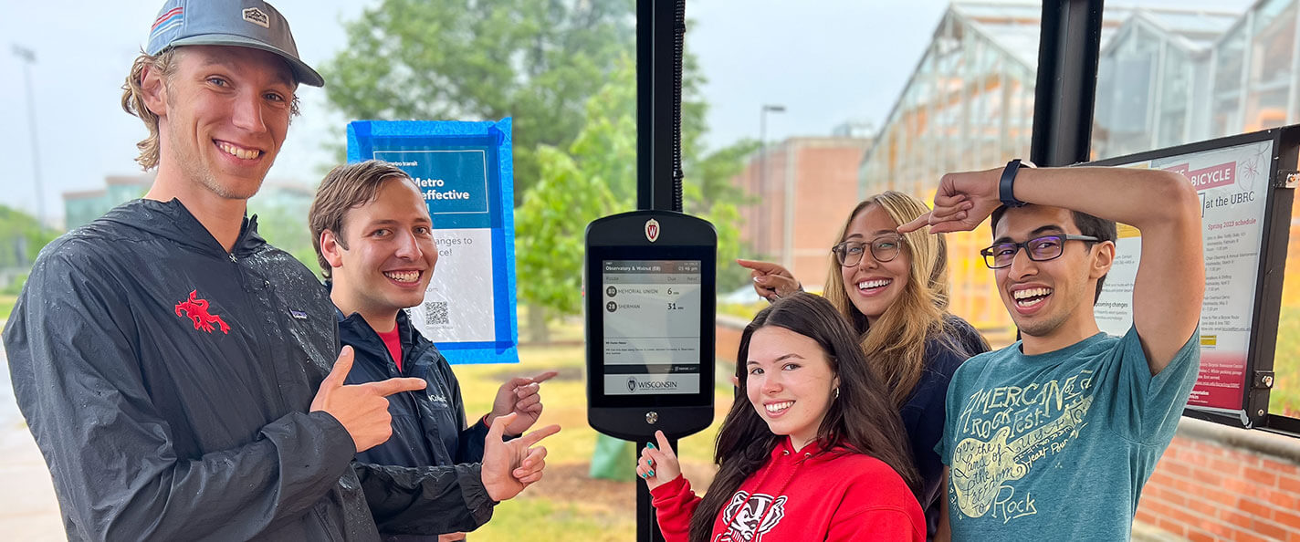 Students pose in front of a solar-powered bus schedule panel.