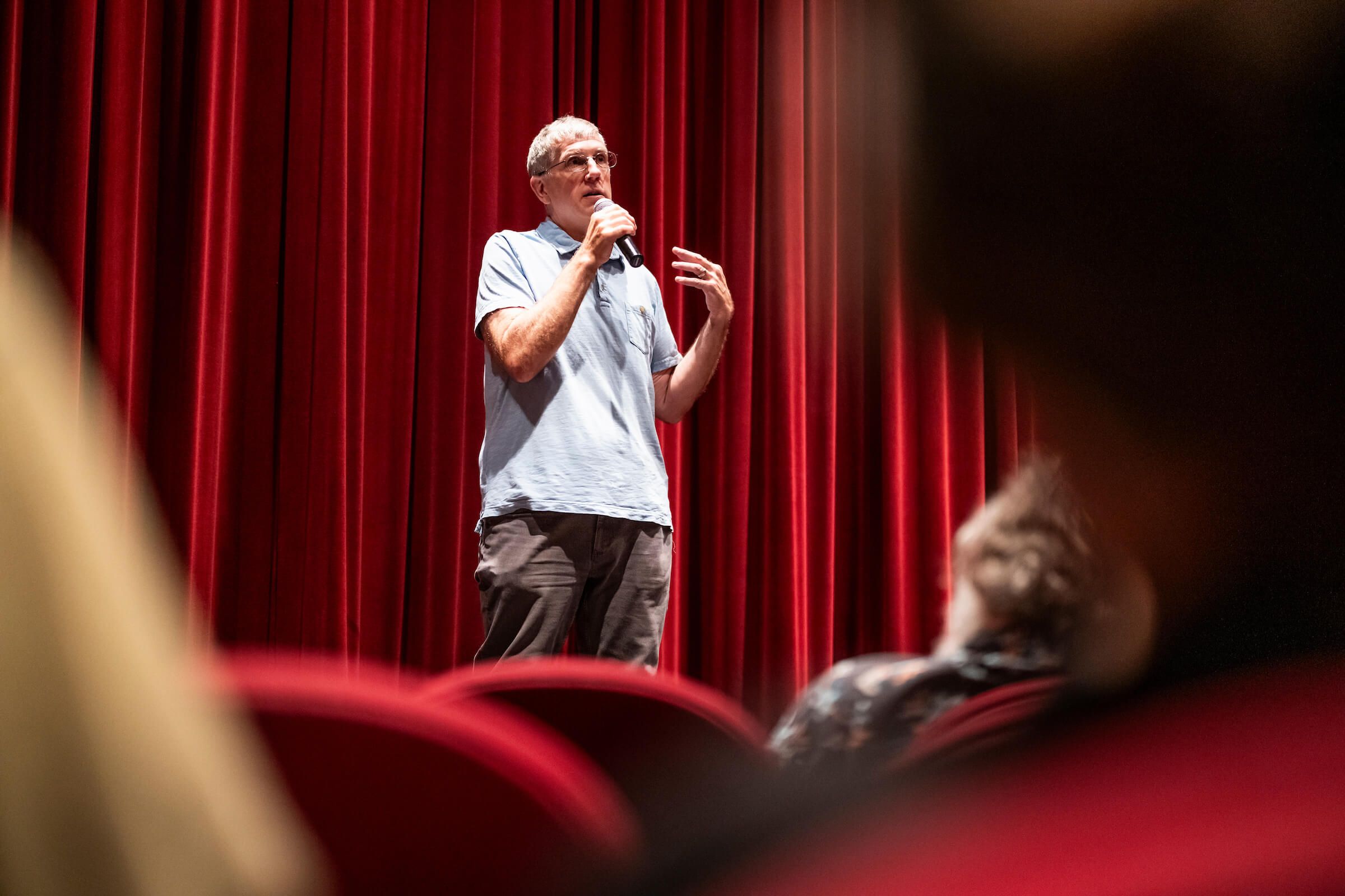 A man wearing a light blue, short sleeve polo shirt and brown slacks is standing in front of a red curtain and holding a microphone. He appears to be addressing an audience. There are red seatbacks and blurred heads of audience members in the foreground.