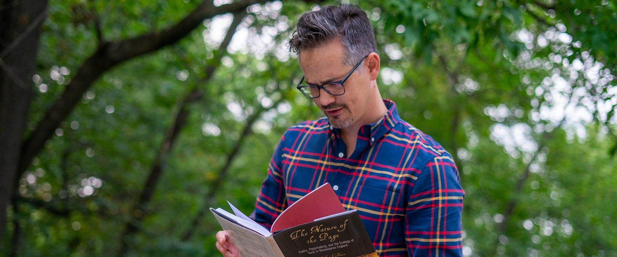 A man in black glasses and a blue and red flannel reads a book. He is set against a background of treetops.