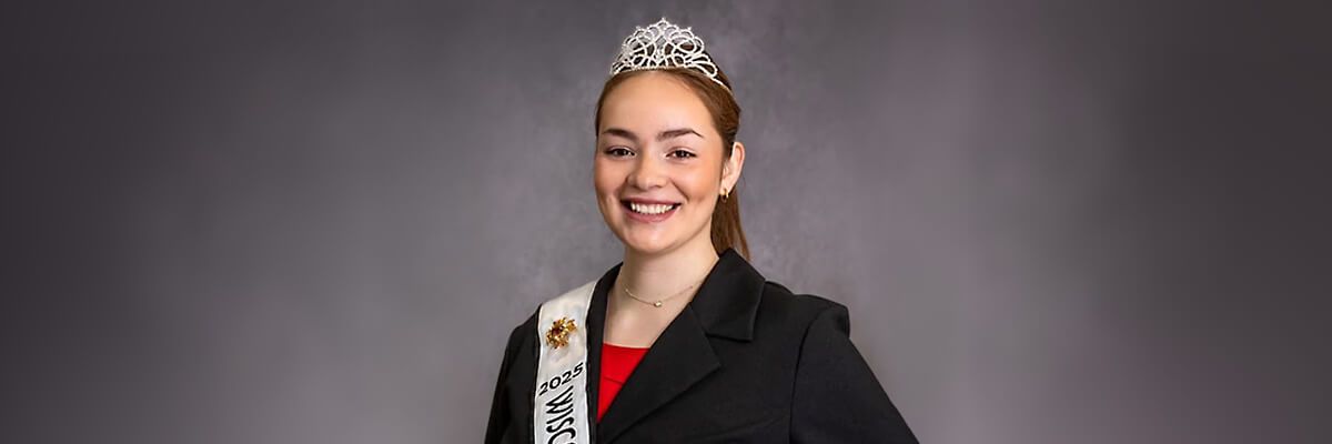 An official portrait of a woman set against a grey backdrop. The woman has dark red hair pulled back in a ponytail and is wearing a small, silver tiara. She is wearing a black blazer over a red top. Over the blazer, she wears a white sash on which a bee emblem and the words "2025 Wisc" are visible.