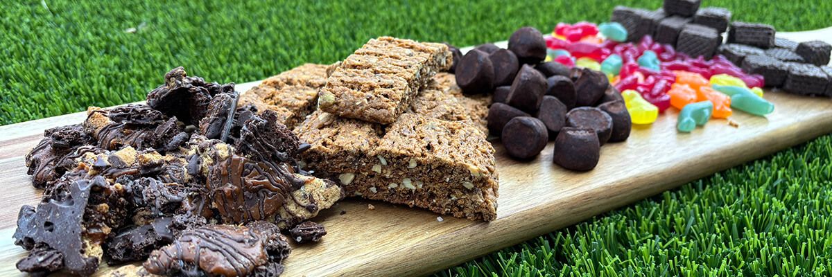 An assortment of treats, including chocolate, rainbow-colored gummy candies, and granola bars, are displayed on a rectangular wooden board sitting on artificial turfgrass.