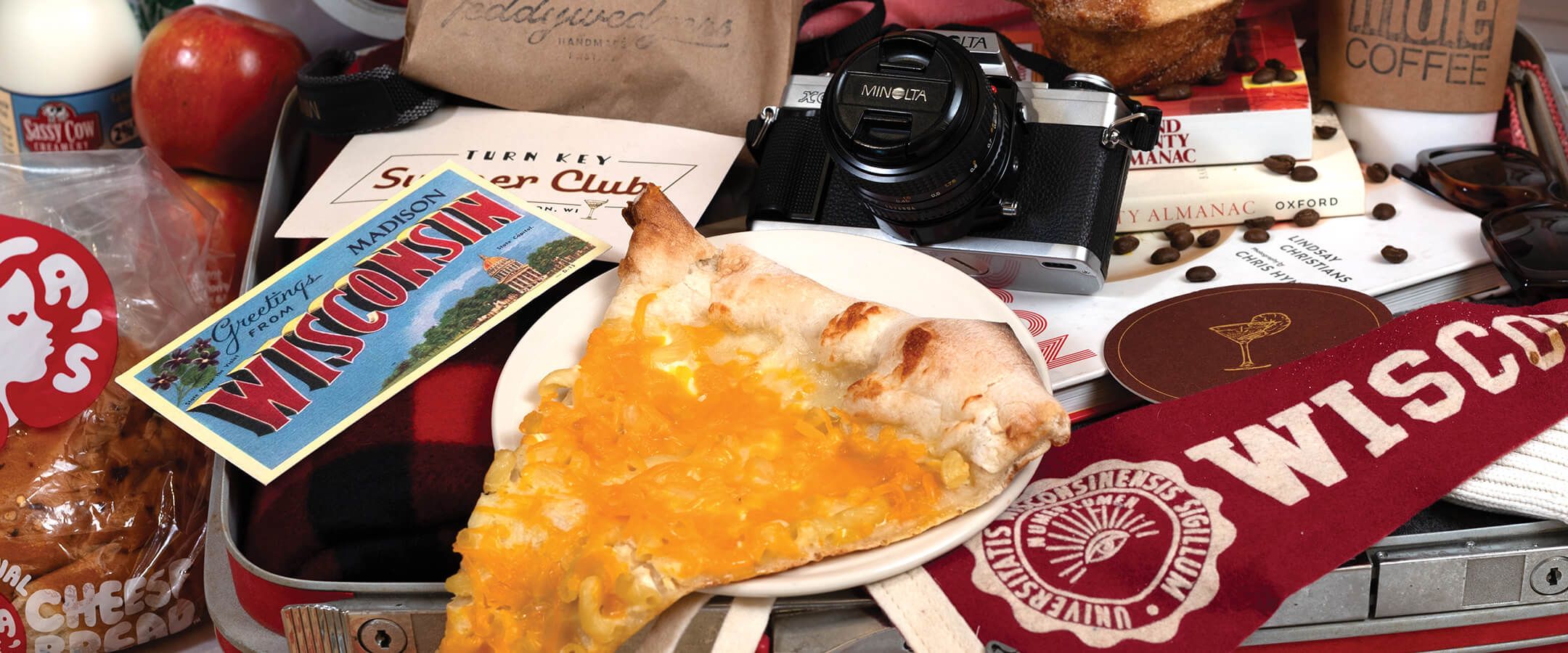 An assortment of items spilling out of a hard-shell suitcase represent Madison, Wisconsin. The collection includes a postcard, a film camera, a red pendant that says "Wisconsin," a slice of pizza on a white plate, an apple, and a loaf of bread in a cellophane bag with a red decal.
