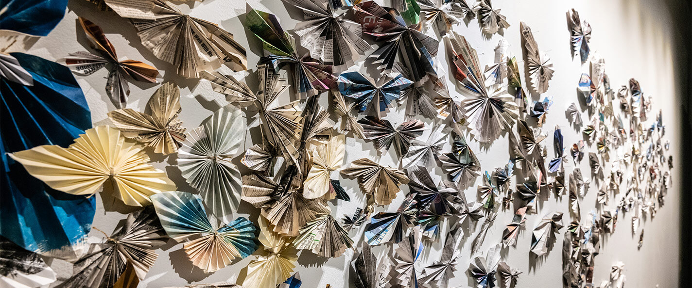 Doznes of folded paper butterflies hung up on the wall of a gallery. 