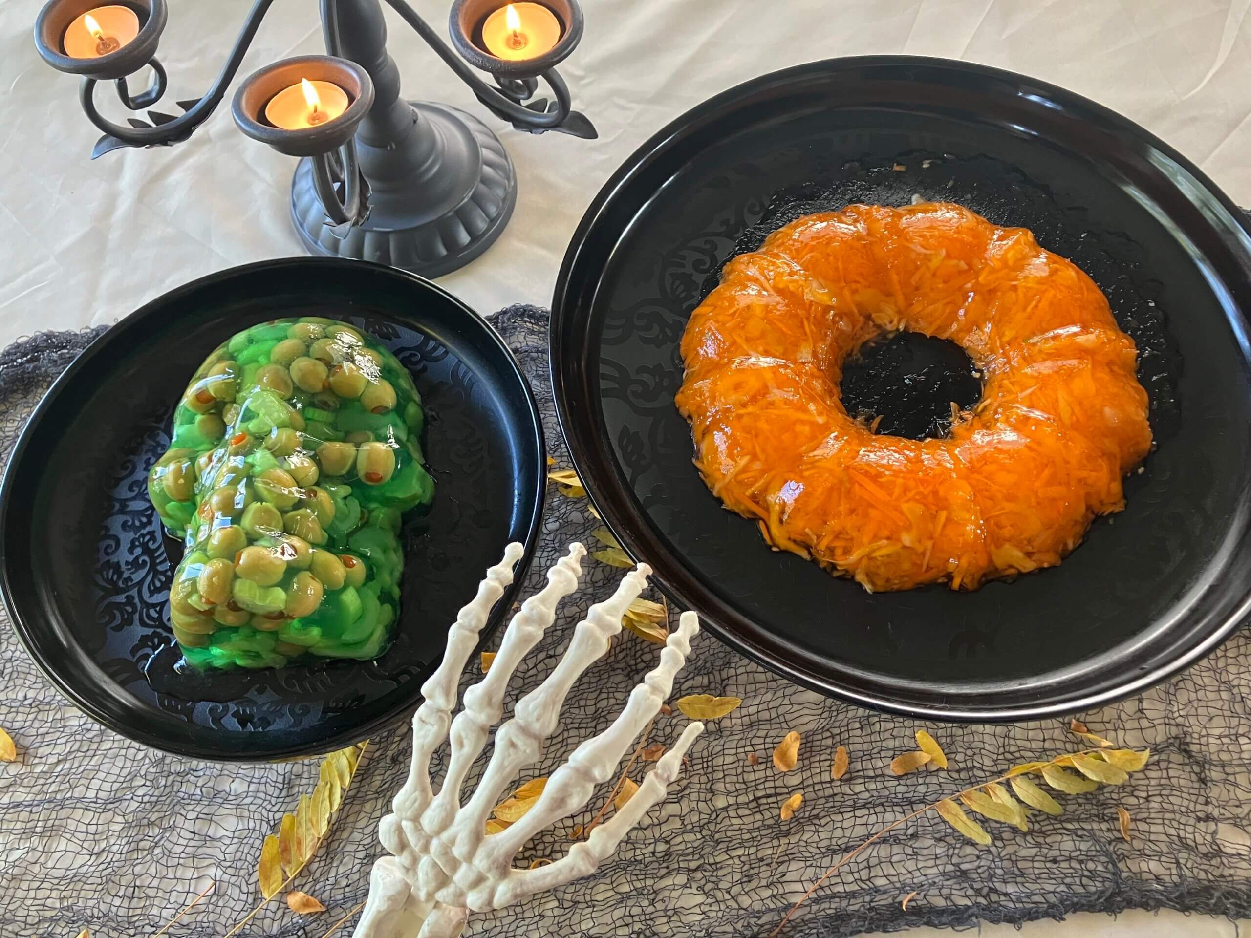 Two Jell-O molds sit side by side on black plates. The one on the left is green, shaped like a skull, and contains green olives. The one on the right is orange, shaped like a ring, and contains flecks of grated carrot and cabbage. In the background is a grey candelabra with orange tea lights. In the foreground is a skeleton hand and a sprig of fall foliage.
