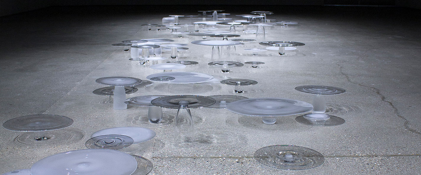 Glass discs sit atop oblong glass vessels on a stone floor. They are lit from above.