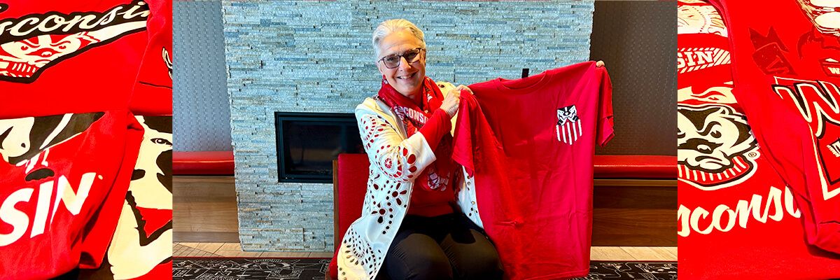 A woman with white hair and glasses wearing a white, crocheted cardigan over a red shirt sits on a red chair in front of a white stone fireplace. She is smiling at the camera and holding a red t-shirt up and to the right. She is flanked by two close-up shots of more red tshirts, each with a different design.