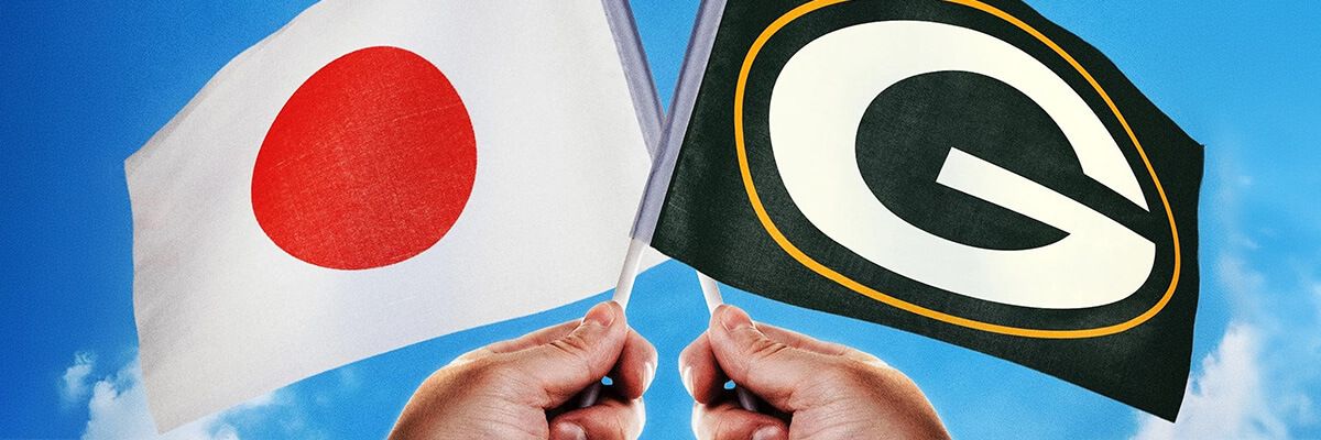 Two hands are crossing two flags against the backdrop of a blue sky. One flag is white with a red circle in the middle (the Japanese flag), and one flag is dark green with a large, white 'G' surrounded by a thin, gold line (Green Bay Packers flag).