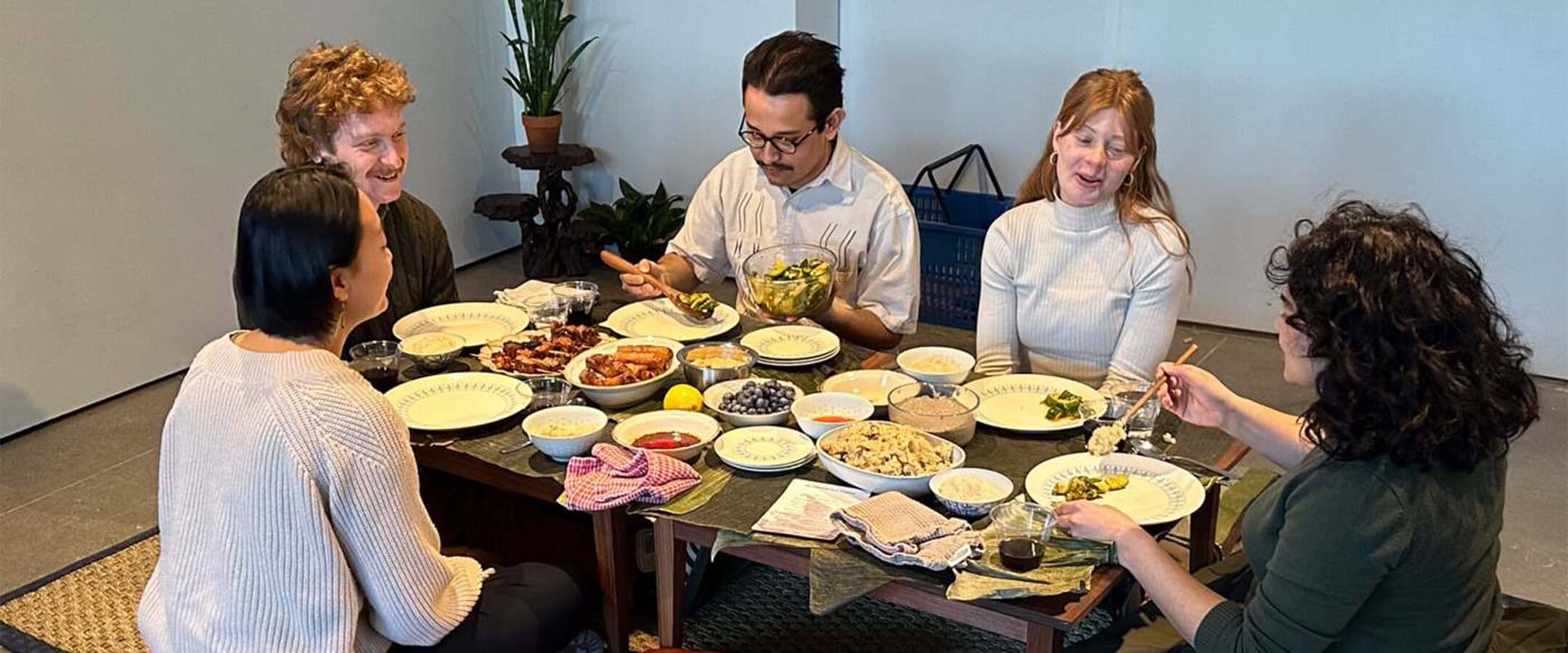 Five adults sit around a table low to the floor in an art gallery eating Filipino food. 