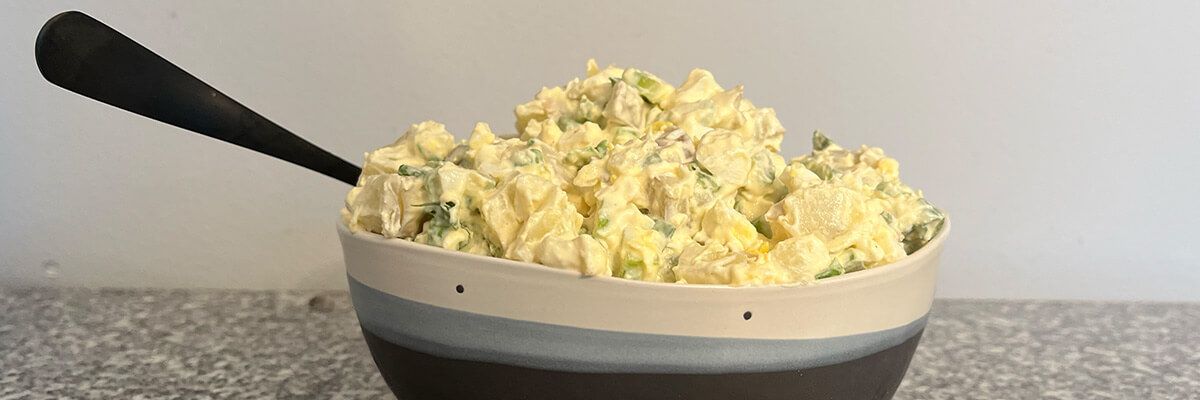 An earthenwear bowl with descending white, light blue, and dark blue stripes sits on a marled white and black countertop. It is full of yellow potato salad. There is a black spoon handle sticking out of the left side of the mound of potato salad.